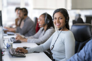 Portrait of smiling businesswoman wearing headset while using computer. Confident female operator is working with colleagues at desk. They are in call center.